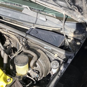 May include: An open car hood revealing engine components. Visible are a black rectangular air intake box, brake fluid reservoir, and various hoses and wires. The hood's interior is black, with a windshield wiper assembly at the top.