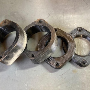 May include: Four dark metal rings with a square flange and circular openings. The rings have a textured surface and appear to be used parts. The objects are arranged on a light gray surface.