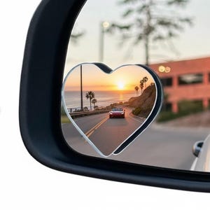 以下が含まれることがあります： 車のサイドミラーに取り付けられたハート型の死角ミラー。 反射には、夕日に向かって走る赤い車のある海岸道路が映っています。 ヤシの木が道路に並んでいます。