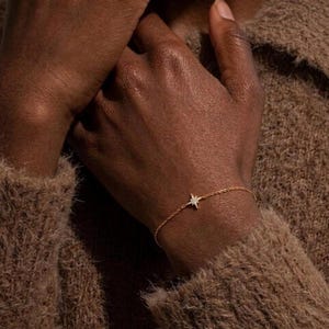 May include: A delicate gold bracelet featuring a starburst charm. The bracelet is worn on a wrist, with the starburst charm centered. The background is a soft, neutral color, complementing the gold of the bracelet.