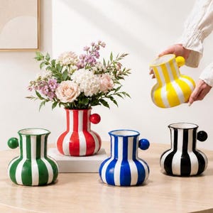 Può includere: Una collezione di vasi in ceramica con strisce verticali e una piccola maniglia sferica sul lato. I vasi sono disponibili in verde, rosso, blu, nero e giallo. Un vaso rosso contiene un bouquet di fiori rosa, bianchi e viola.
