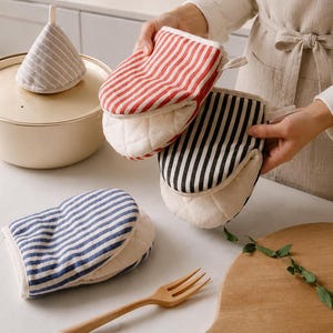 May include: Three quilted oven mitts featuring striped patterns in red, black, and blue, alongside a matching cone-shaped pot lid handle cover. The mitts have a white interior lining and a soft fabric texture, designed for handling hot kitchen cookware.