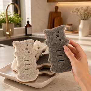 May include: A collection of cat-shaped kitchen sponges in various shades of gray and white. The sponges have a textured surface and are stacked on a white rectangular plate. One sponge is being held up by a hand.