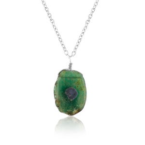 Può includere: Una collana a catena in metallo color argento con un ciondolo composto da una pietra verde grezza di forma irregolare. La pietra presenta un'inclusione circolare scura al centro ed è fissata alla catena da un gancio in filo metallico.