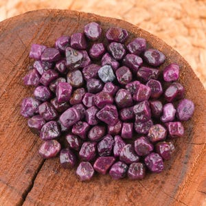 May include: A collection of raw, uncut ruby gemstones in varying shades of deep red and purple. The gemstones are displayed on a rustic, wooden surface, showcasing their natural, irregular shapes and textures. The lighting highlights the stones' luster.