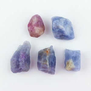 May include: Five raw gemstones in shades of purple, blue, and pink are arranged on a white surface. The stones have a rough, natural texture and vary in size and shape. The colors range from deep violet to pale blue and pink.