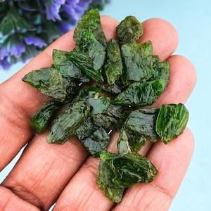 Pode incluir: Um punhado de pedras preciosas verdes brutas e ásperas. As pedras têm uma cor verde esmeralda profunda e uma aparência cristalina e texturizada. As pedras preciosas são seguradas em uma mão aberta contra um fundo azul claro.