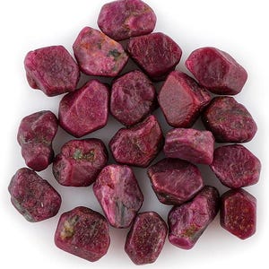 Op de afbeelding: Een verzameling ruwe robijn edelstenen, elk met een dieprode tint en onregelmatige, natuurlijke vormen. De stenen variëren in grootte en hebben een enigszins ruwe textuur, met enkele groene hints.