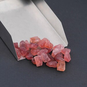 Puede incluir: Una pala de acero inoxidable que contiene una colecci&oacute;n de peque&ntilde;os fragmentos de piedras preciosas en bruto, transl&uacute;cidas y de color rojo rosado. Las piedras tienen formas angulares irregulares y profundidades de color variables.