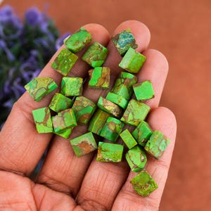 May include: A collection of bright green turquoise gemstone beads with brown veining. The beads are in various irregular shapes, with a mix of angular and rounded edges. The gemstones are held in a hand, with a blurred background.
