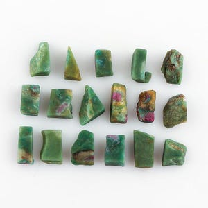 Op de afbeelding: Een verzameling ruwe, ongeslepen edelstenen in groentinten, met hints van rood en bruin. De stenen variëren in vorm, waaronder rechthoekige en driehoekige vormen, en worden weergegeven tegen een witte achtergrond.