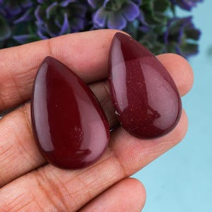 May include: Two teardrop-shaped, polished gemstones in a deep burgundy color. The stones have a smooth, glossy surface and are displayed against a light blue background. The gemstones are likely for jewelry making or crafting.
