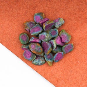 May include: A collection of small, raw, tumbled stones featuring a vibrant magenta-pink center surrounded by a textured, mottled green and grey outer layer. These irregular, multi-faceted mineral pieces are clustered together in a dense pile.