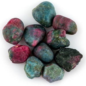 Op de afbeelding: Een verzameling getrommelde robijn-in-zoisiet stenen. De stenen vertonen een mix van kleuren, waaronder diep rood, groen en blauw. De stenen hebben een ruwe, natuurlijke textuur en variëren in grootte en vorm. De achtergrond is wit.