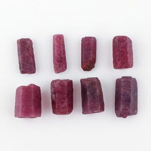 Peut inclure: Huit pierres précieuses de rubis brutes, de couleur rouge-violet profond. Les pierres sont de forme rectangulaire avec des bords facettés, présentant des textures naturelles. Elles sont disposées sur un fond blanc.