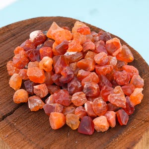 May include: A pile of raw, tumbled carnelian gemstones in shades of orange and red. The gemstones are displayed on a rustic wooden surface against a light blue background. The stones vary in size and shape, with a natural, unpolished appearance.