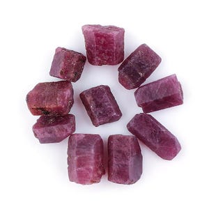 Może przedstawiać: Kolekcja surowych kamieni szlachetnych rubinu, każdy o głębokim czerwono-różowym odcieniu. Kamienie różnią się kształtem, z widocznymi fasetami i naturalnymi teksturami. Ułożone są w okrąg na białym tle.