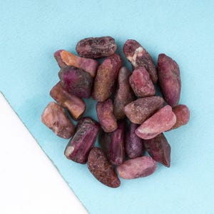Pode incluir: Uma coleção de pedras preciosas de rubi bruto em várias formas e tamanhos. As pedras exibem uma gama de cores, do bordô profundo ao rosa pálido, com algumas exibindo tons mistos. As pedras preciosas estão espalhadas sobre uma superfície azul clara.
