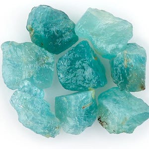 Op de afbeelding: Een verzameling ruwe aquamarijn edelstenen. De onbewerkte kristallen vertonen een lichtblauwe tot groenblauwe kleur en onregelmatige vormen. De doorschijnende stenen variëren in grootte en tonen hun natuurlijke, ongeraffineerde textuur. Geschikt voor sieradenontwerp of als verzamelobjecten.