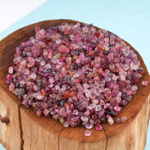 Può includere: Una ciotola di legno piena di una collezione di piccole pietre preziose grezze. Le pietre preziose sono principalmente sfumature di rosa, rosso e viola, con alcune variazioni più scure e chiare. La ciotola è di legno marrone chiaro.