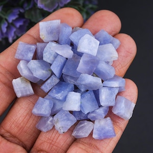 Könnte beinhalten: Eine Handvoll roher, hellblau-violetter Tansanit-Edelsteine. Die rohen, ungeschliffenen Steine variieren in Größe und Form und weisen eine natürliche, kristalline Textur auf. Die Edelsteine werden in einer offenen Hand vor dunklem Hintergrund gehalten.