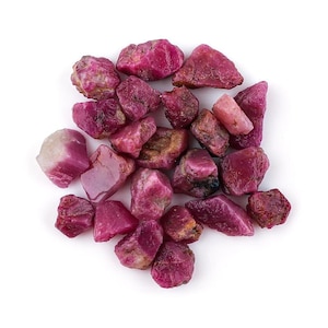 Puede incluir: Una colección de piedras preciosas de rubí en bruto de varias formas y tamaños. Las piedras muestran una gama de tonos rosados y rojizos, algunas con una calidad translúcida. Las piedras preciosas están dispersas sobre un fondo blanco.