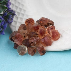 Puede incluir: Un conjunto de gemas en bruto, sin cortar, en tonos marrones y ámbar. Los cristales están agrupados sobre una superficie blanca texturizada, con un fondo azul. Las gemas varían en tamaño y forma, mostrando su textura natural y rugosa.
