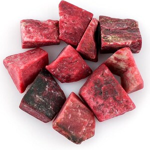 Peut inclure: Un ensemble de pierres précieuses brutes de couleur rouge. Les pierres varient en forme, certaines apparaissant rectangulaires et d'autres triangulaires. La couleur varie du rouge profond et riche aux nuances plus claires avec quelques inclusions sombres. Les pierres sont disposées sur une surface blanche.