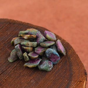 May include: A pile of rough-cut ruby gemstones with a mix of green, blue, and pink hues. The gemstones are displayed on a rustic, dark brown wooden surface, with a reddish-orange background.