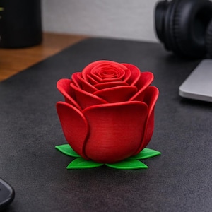 Puede incluir: Una vibrante escultura de rosa roja con hojas verdes, sobre una superficie oscura. Los pétalos detallados y el diseño realista crean un impacto visual llamativo. Un acento floral decorativo para la decoración del hogar o para regalar.