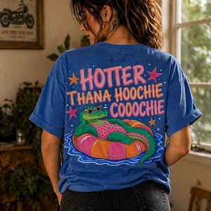 Puede incluir: Camiseta azul con el texto "HOTTTER THANA HOOCHIE COOOCHIE" y un gráfico de un caimán verde con gafas de sol flotando en una cámara de aire rosa y naranja. La camiseta tiene las mangas enrolladas.