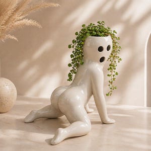May include: A white ceramic planter in the shape of a crawling figure with three facial openings and a round base. The planter is filled with green trailing plants. Decorative vase and sphere are in the background.
