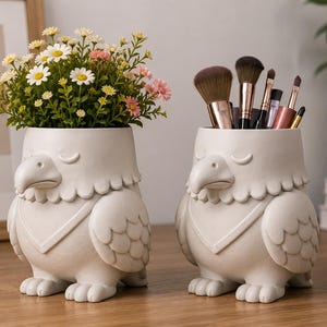 May include: Two off-white, eagle-shaped ceramic containers. One holds a bouquet of daisies and other flowers, the other holds makeup brushes. The containers have a detailed design, including a beak, wings, and feet.