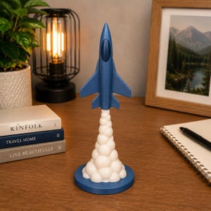 Pode incluir: Estatueta de foguete azul com detalhes de fumaça branca, sobre uma base azul. O foguete tem asas e um nariz pontudo. A estatueta é um item decorativo, possivelmente um adorno de mesa.