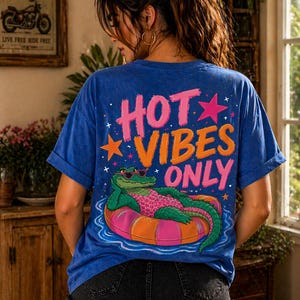 Può includere: Maglietta blu con la scritta "HOT VIBES ONLY" in rosa e arancione, con una grafica di un alligatore con occhiali da sole su un salvagente. La maglietta è abbinata a pantaloncini di jeans neri.