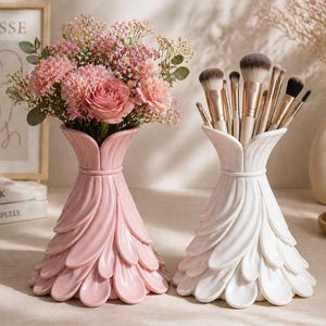 May include: Two decorative vases, one pink and one white, with a petal-like design. The pink vase holds a bouquet of pink flowers, while the white vase contains makeup brushes. The vases are on a light-colored surface.