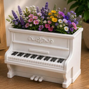 Op de afbeelding: Een witte piano-vormige plantenbak gevuld met een levendig arrangement van paarse lavendel, roze madeliefjes, gele bloemen en witte bloesems. De plantenbak is voorzien van gedetailleerde zwarte en witte toetsen, drie pedalen en decoratieve lijsten.