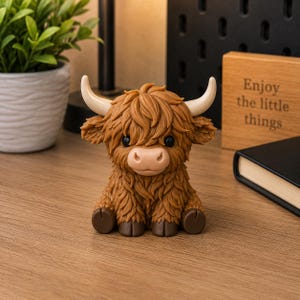 May include: A detailed figurine of a Highland cow, featuring a shaggy brown coat, small black eyes, and curved cream horns. The cow is positioned on a wooden surface. A wooden block with the text "Enjoy the little things" is visible.