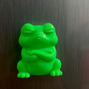 May include: A bright green frog figurine with a smooth, glossy finish. The frog is depicted in a seated position with its eyes closed and its front arms crossed firmly across its chest, creating a stern and unimpressed facial expression.