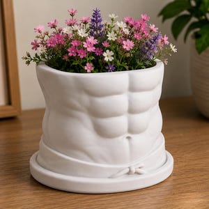 May include: A white ceramic planter shaped like a human torso with defined abdominal muscles. The planter sits on a matching circular base and is filled with a vibrant arrangement of small pink, white, and purple flowers with green foliage.
