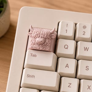 May include: A custom keyboard keycap featuring a Highland cow design in a light pink color. The keycap is installed on a beige keyboard, with the "Tab" key visible. The keyboard is on a wooden surface, with a potted plant in the background.