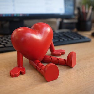 May include: A red heart-shaped figurine with articulated legs and feet, sitting on a wooden desk. The figurine has a glossy finish and is positioned in front of a computer keyboard and monitor. The desk also has a mouse and pen holder.
