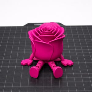 Könnte beinhalten: Eine leuchtend magentafarbene Rosenfigur mit einem skurrilen Design. Die Rose hat eine detaillierte Blütenblattstruktur und sitzt mit angebrachten Beinen und Armen. Die Figur steht auf einer schwarzen Gitteroberfläche.