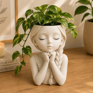 May include: A cream-colored resin planter shaped like a person with closed eyes, resting their chin on clasped hands. The figure features a side braid and a scalloped neckline. A green and yellow variegated pothos plant grows from the top.