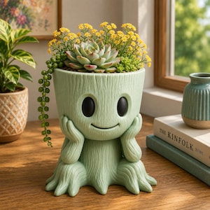 Puede incluir: Una maceta de cerámica de color verde claro con forma de personaje arbóreo, con grandes ojos negros y una boca sonriente. La maceta contiene varias suculentas pequeñas, incluyendo una planta en forma de roseta y una planta colgante.