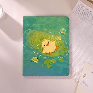 May include: A green and blue notebook featuring a cartoon duckling floating in water with a frog and lily pads. The duckling is yellow with an orange beak and feet. A bubble floats above the duckling.