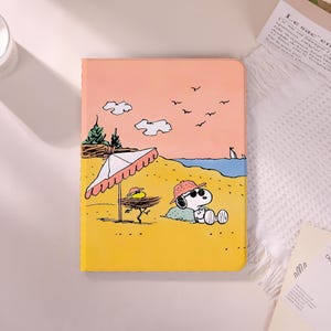 Op de afbeelding: Een gele en roze tablethoes met een strandscène-illustratie. Het ontwerp toont Snoopy met zonnebril en hoed, een parasol, wolken, vogels en de oceaan.