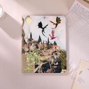 May include: A fantasy-themed journal featuring a castle illustration with flying dragons in black, green, and pink. The cover also includes text from a book page and floral accents, creating a whimsical design.
