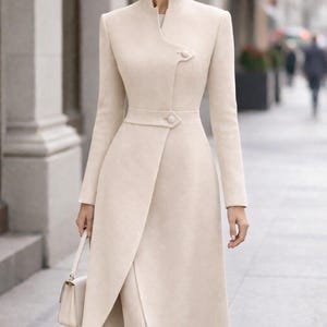 May include: A cream-colored, long coat with a wrap-style front and a high collar. The coat features a belted waist and button details. The overall design is elegant and tailored, suitable for formal occasions. The coat is paired with matching pants and a small handbag.