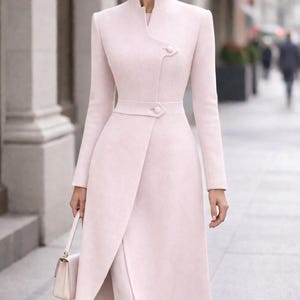 May include: A light pink, long coat with a high collar and a wrap-style front. The coat features a button closure at the waist and a matching belt. The coat is paired with matching pants and a small, light pink handbag. The coat is a sophisticated, elegant design.